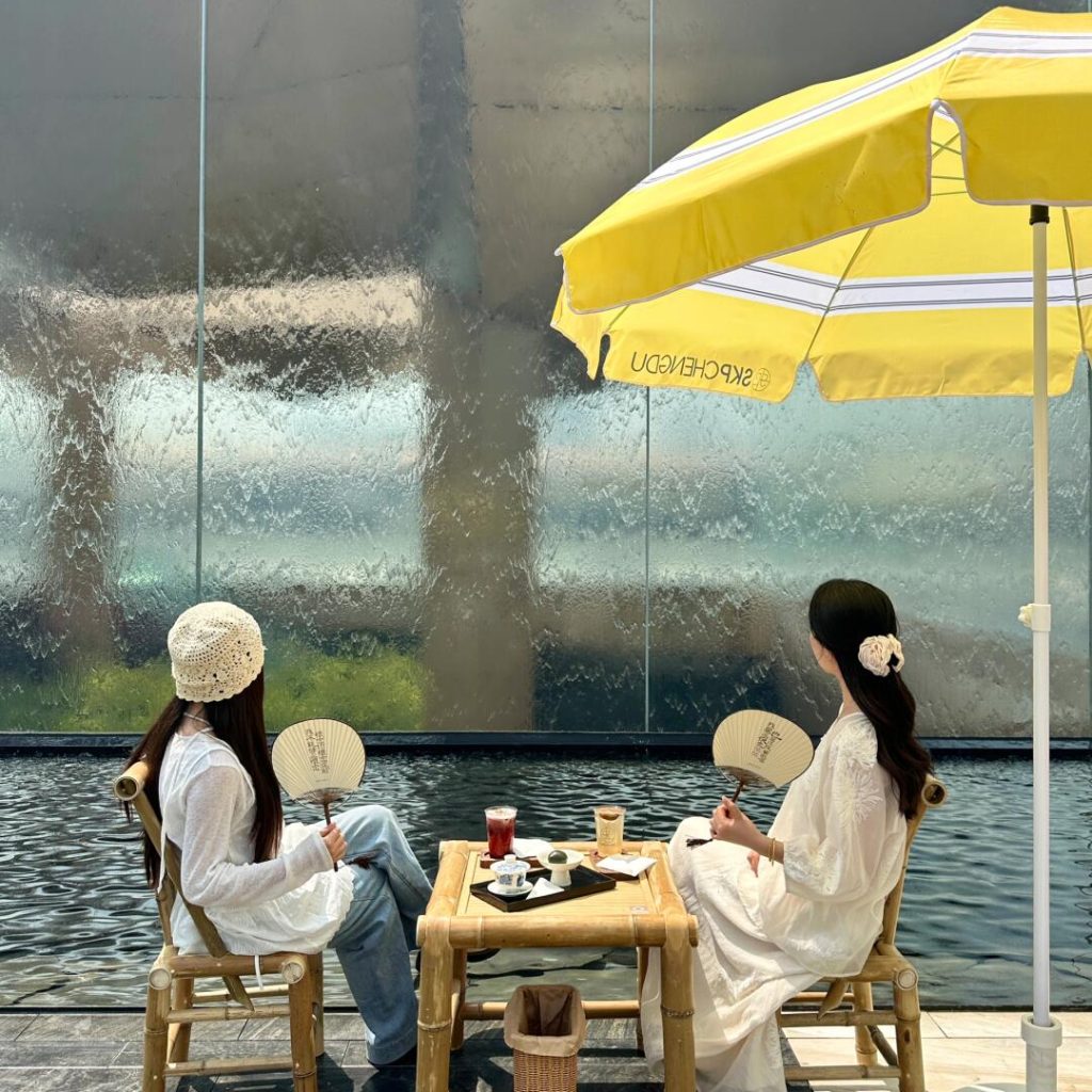 Two women enjoying tea beside a modern water-wall installation at SKP Chengdu, showcasing contemporary Chengdu teahouse culture.